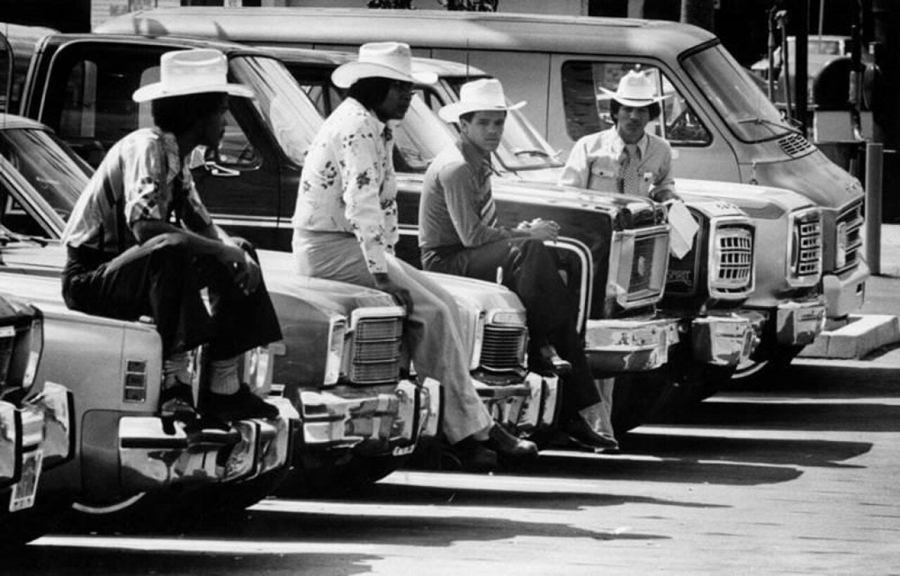 Four car salesman hanging around cars with cowboy hats on