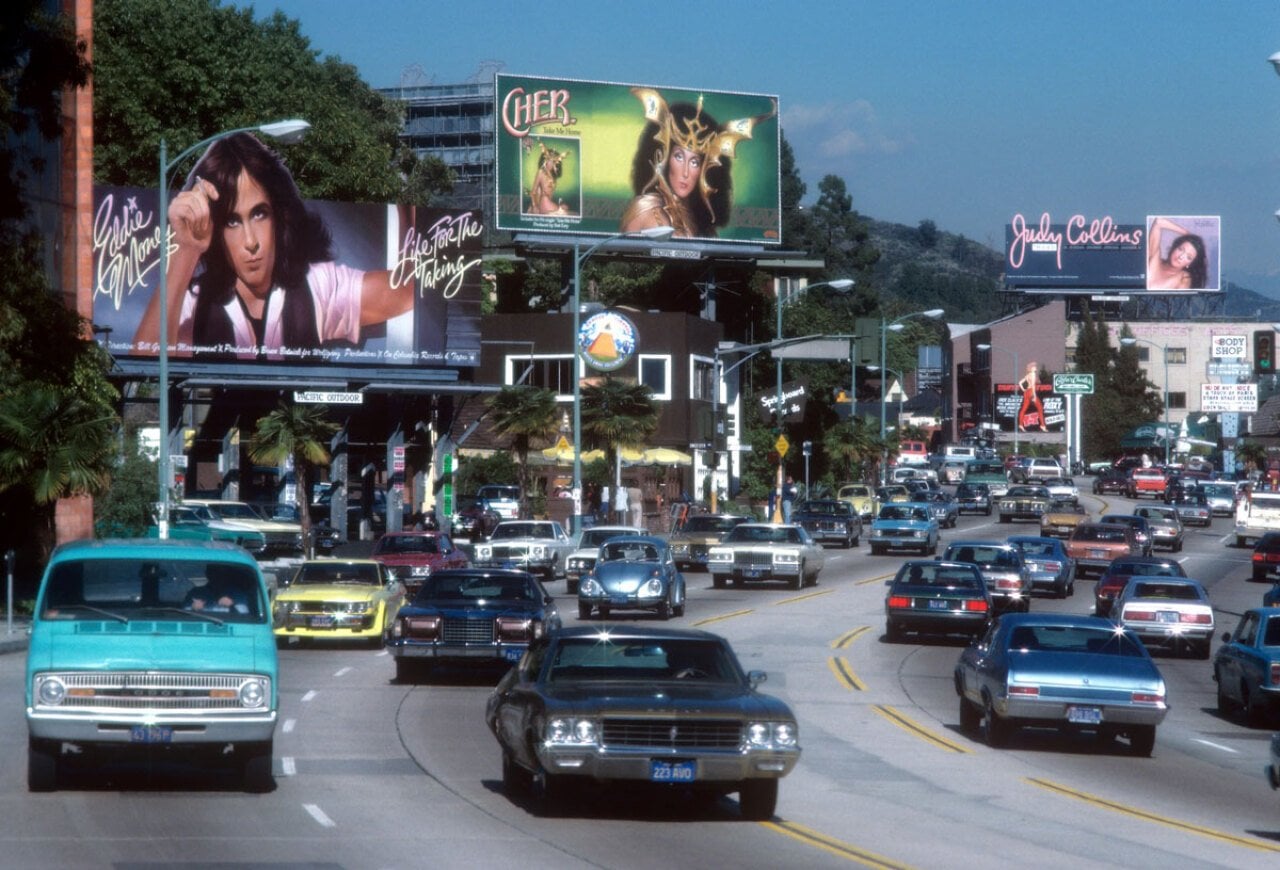 Cars driving down the sunset strip in LA with billboards for Eddie Money, Cher, and Judy Collins