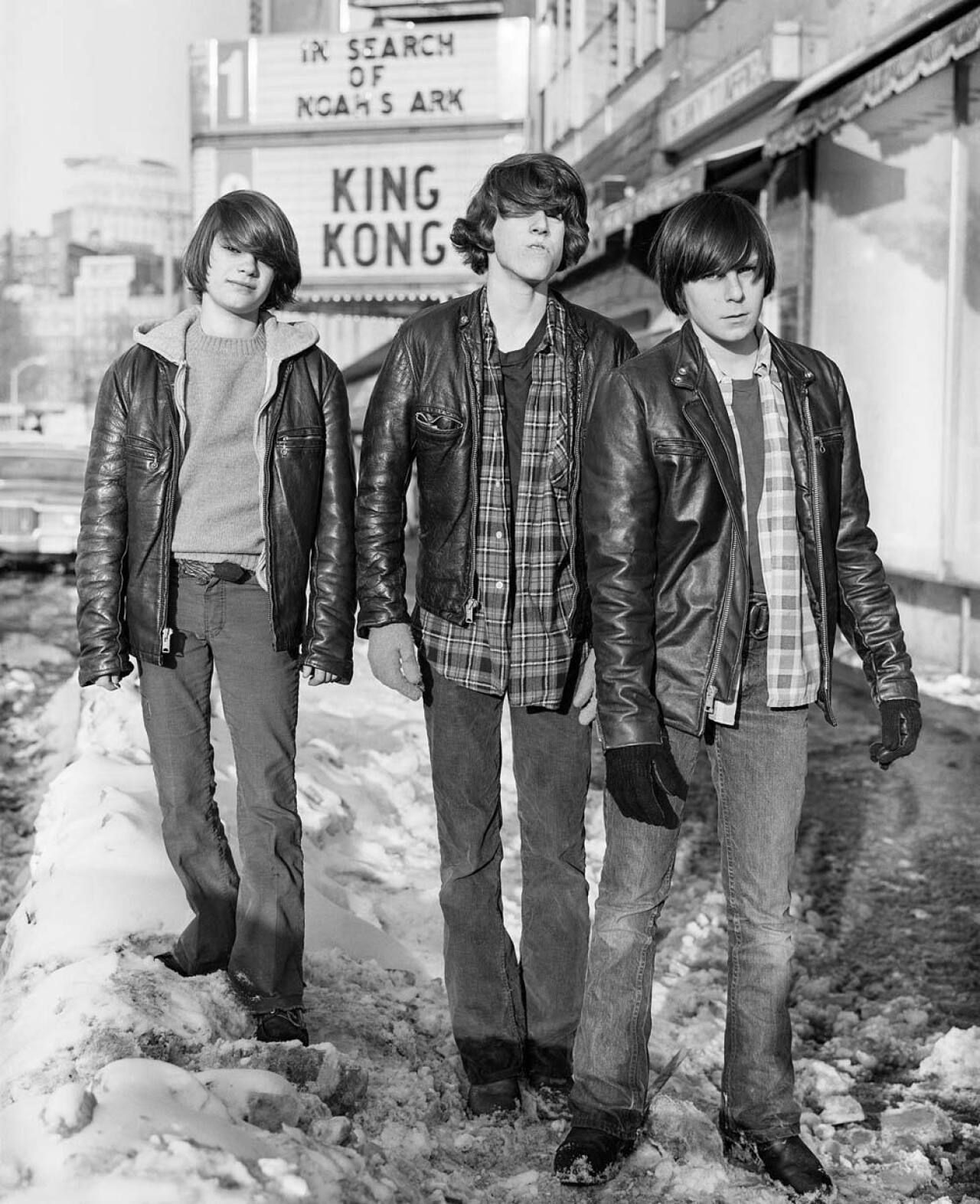 Black-and-white photo of three teenage boys in leather jackets walking down a snowy Boylston Street in Boston. All three have bowl-cut hair and wear plaid shirts underneath their jackets. A movie theater marquee behind them reads 
