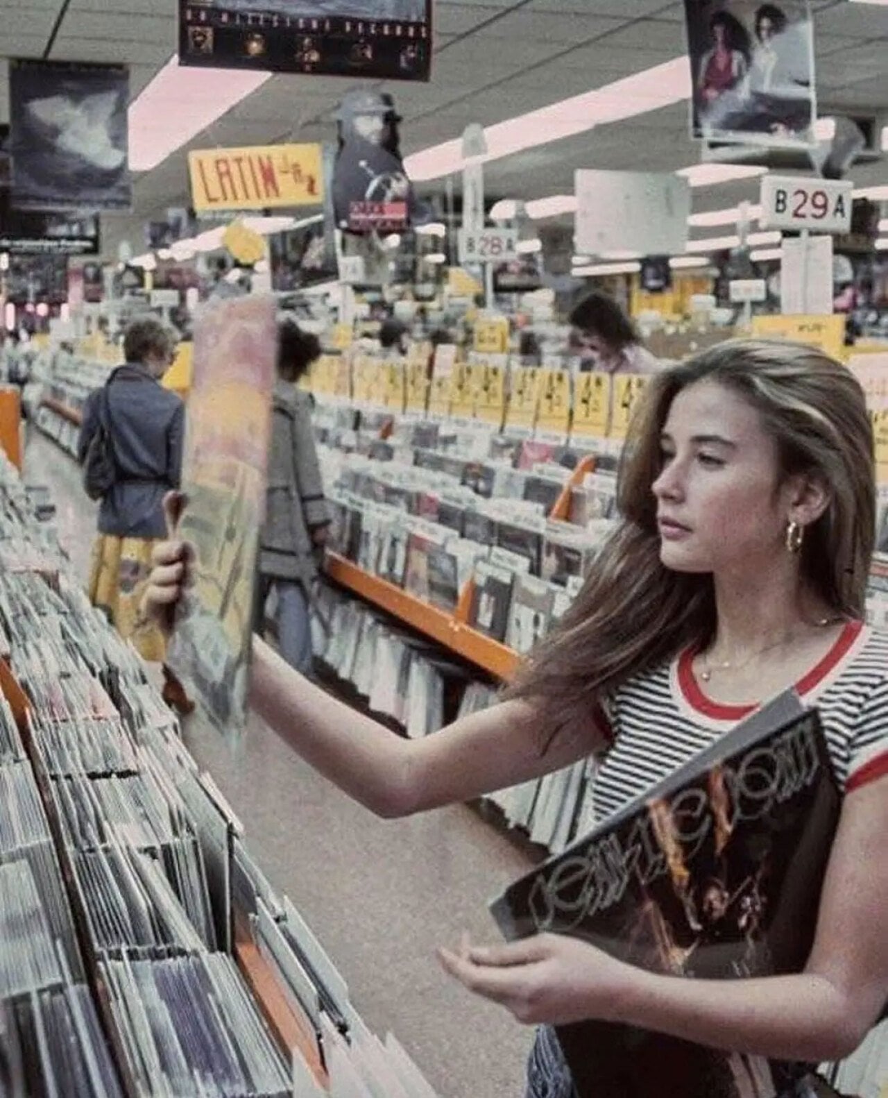 Color photo of a teenage Demi Moore browsing vinyl records at Tower Records on the Sunset Strip in Los Angeles, 1977. She wears a striped red-and-white tank top and hoop earrings, and holds a record album while standing in an aisle lined floor to ceiling with LP covers. A 