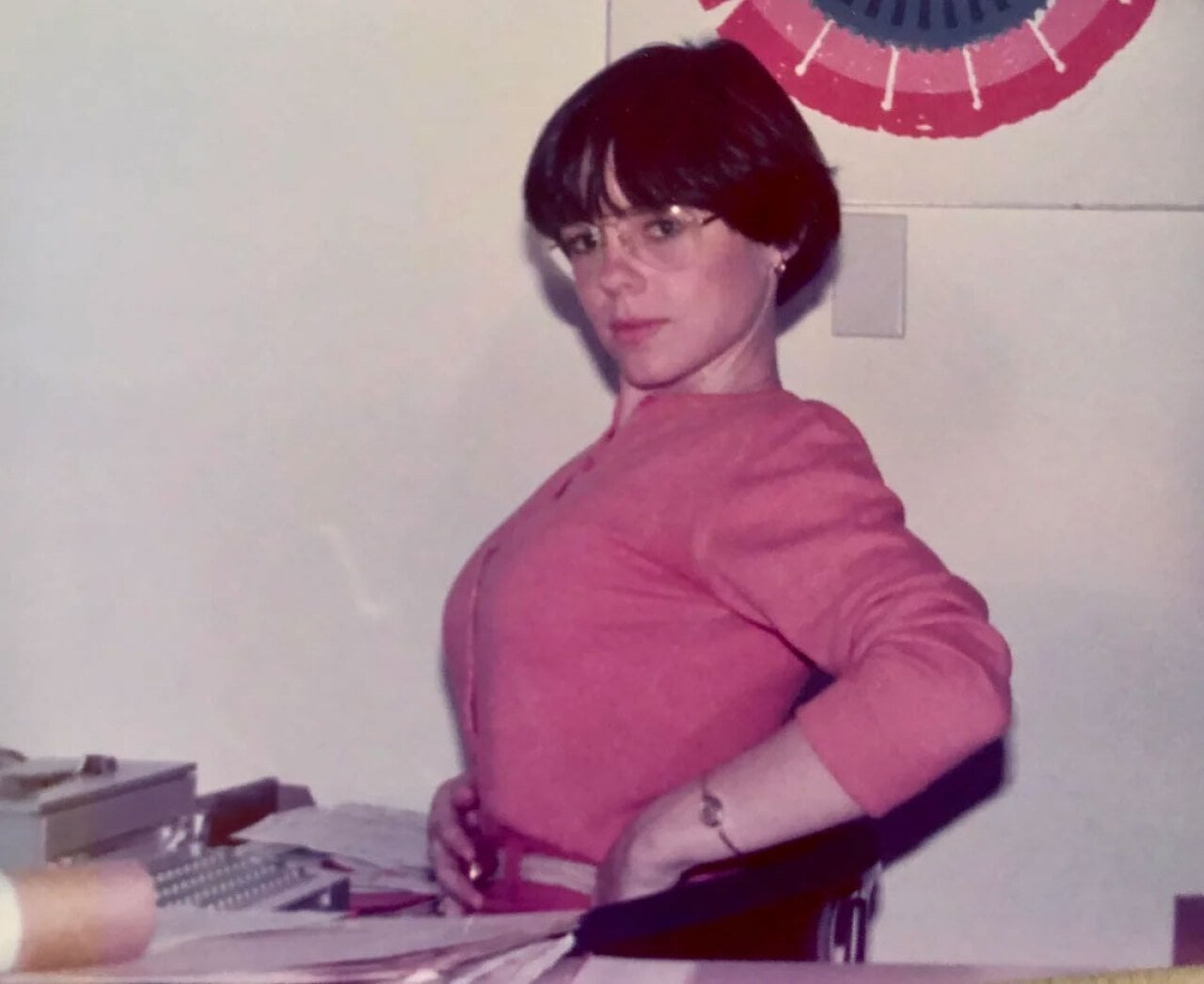 Color photo of a woman seated at an office desk in 1977, looking over her shoulder at the camera. She wears a pink top, wire-rimmed glasses, and a short dark bowl cut. Papers and a typewriter are visible on the desk; a red, white, and blue decorative bunting hangs on the wall behind her.