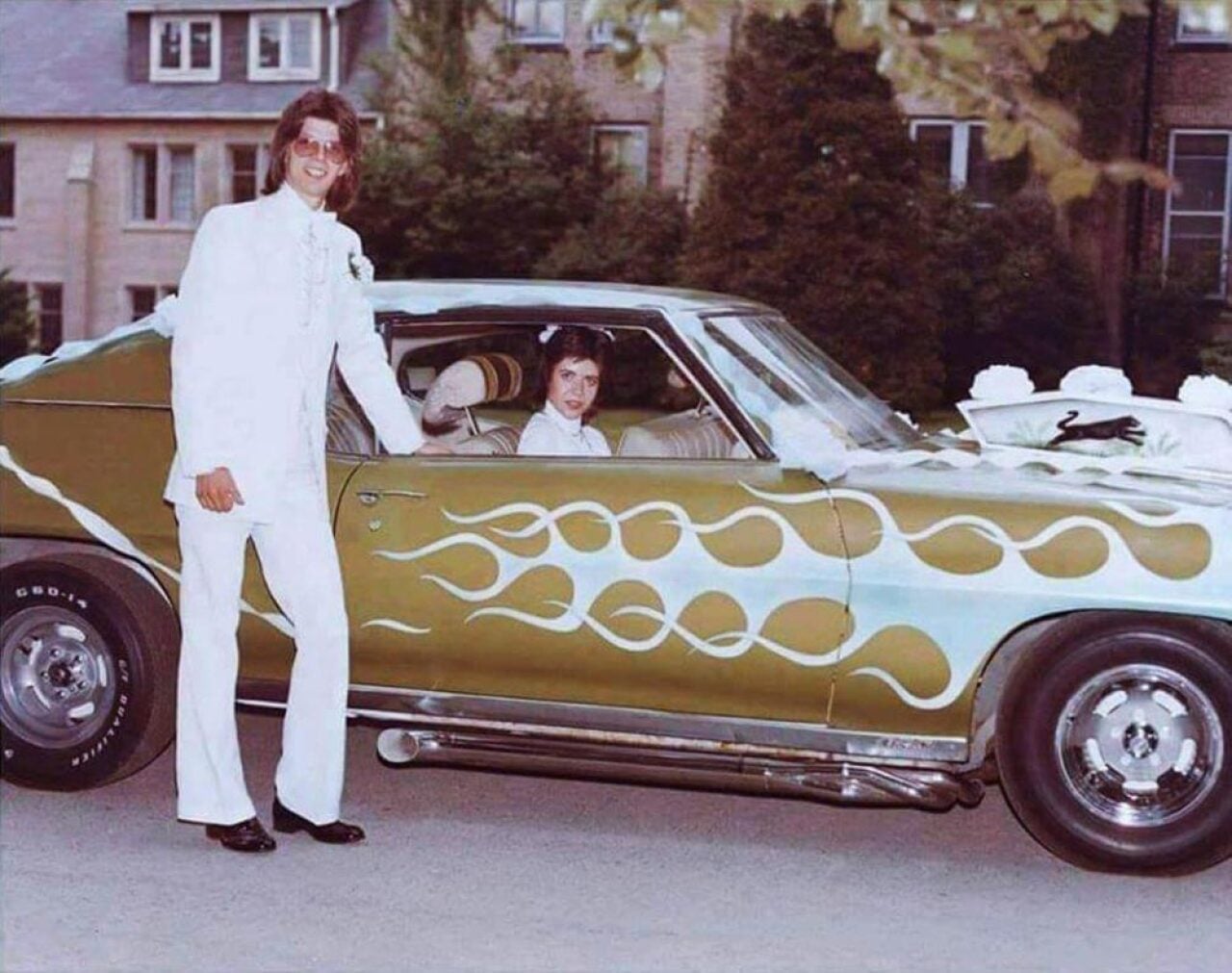 Color photo of a young groom in a white suit and aviator sunglasses standing beside his muscle car, decorated with white floral wedding ribbons. His bride sits in the passenger seat. A brick building and trees are visible in the background.