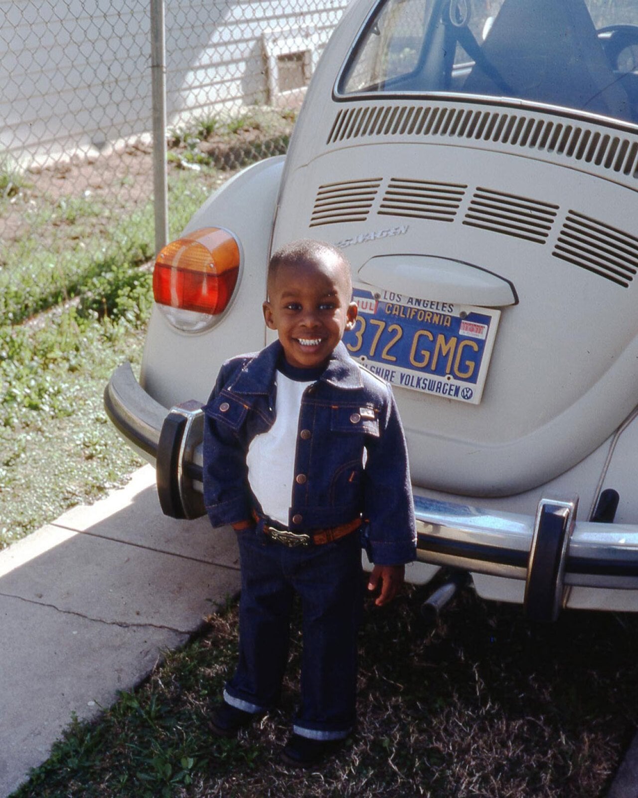 Color photo of a young boy standing in front of a white Volkswagen Beetle in Los Angeles, 1977. He wears a matching dark denim jacket and jeans with a white turtleneck and a brown belt with a large brass buckle, and grins broadly at the camera. A California license plate reading 