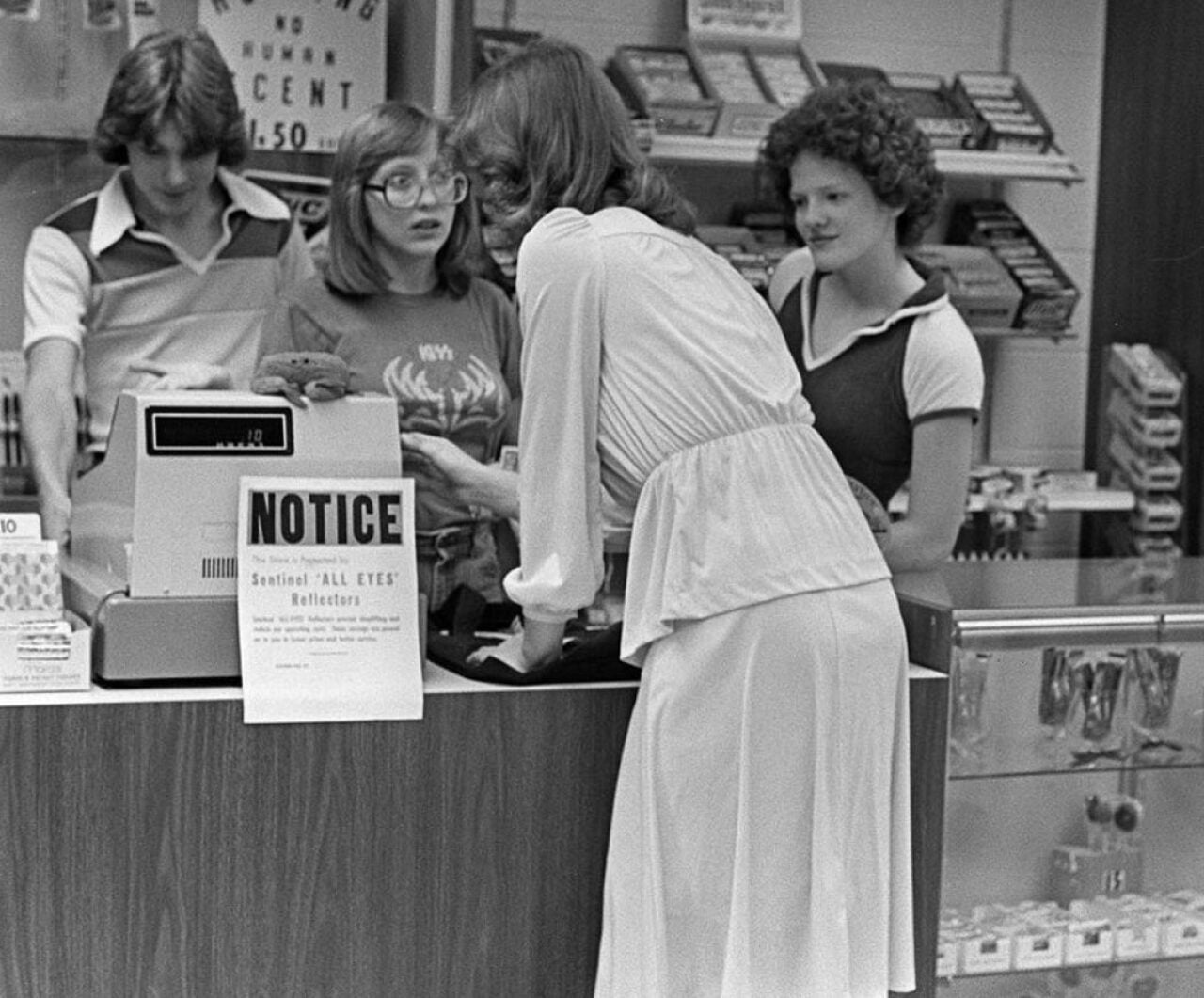 Black-and-white photo of a customer leaning over a retail counter toward two young female cashiers at a store in the Poconos, Pennsylvania, in 1977. The cashier on the left wears glasses and a KISS t-shirt; the cashier on the right looks on with a slight smile. A 