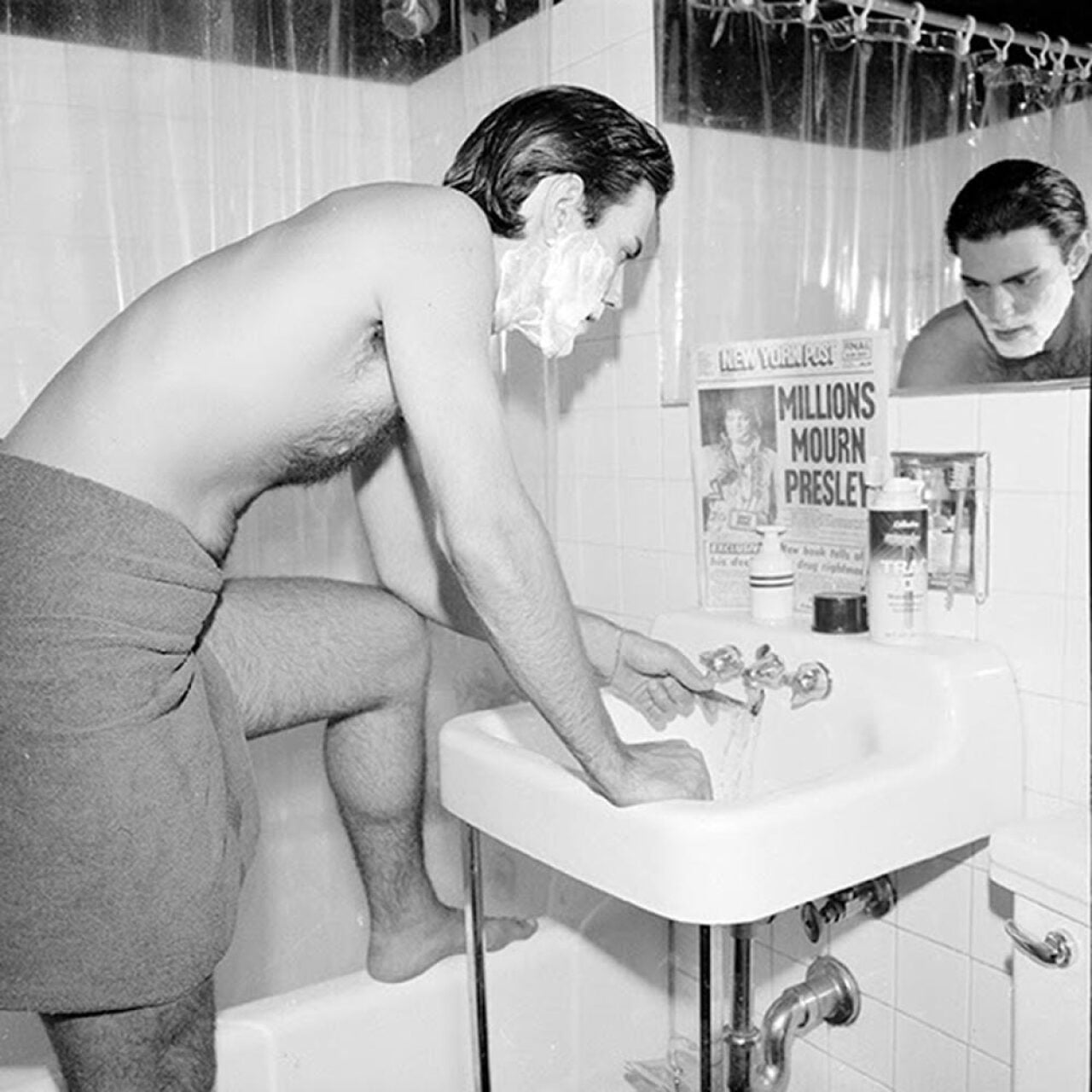 Black-and-white photo of a shirtless man leaning over a bathroom sink to shave, his face covered in shaving cream. Propped against the mirror is a copy of the New York Post with the front page headline reading 