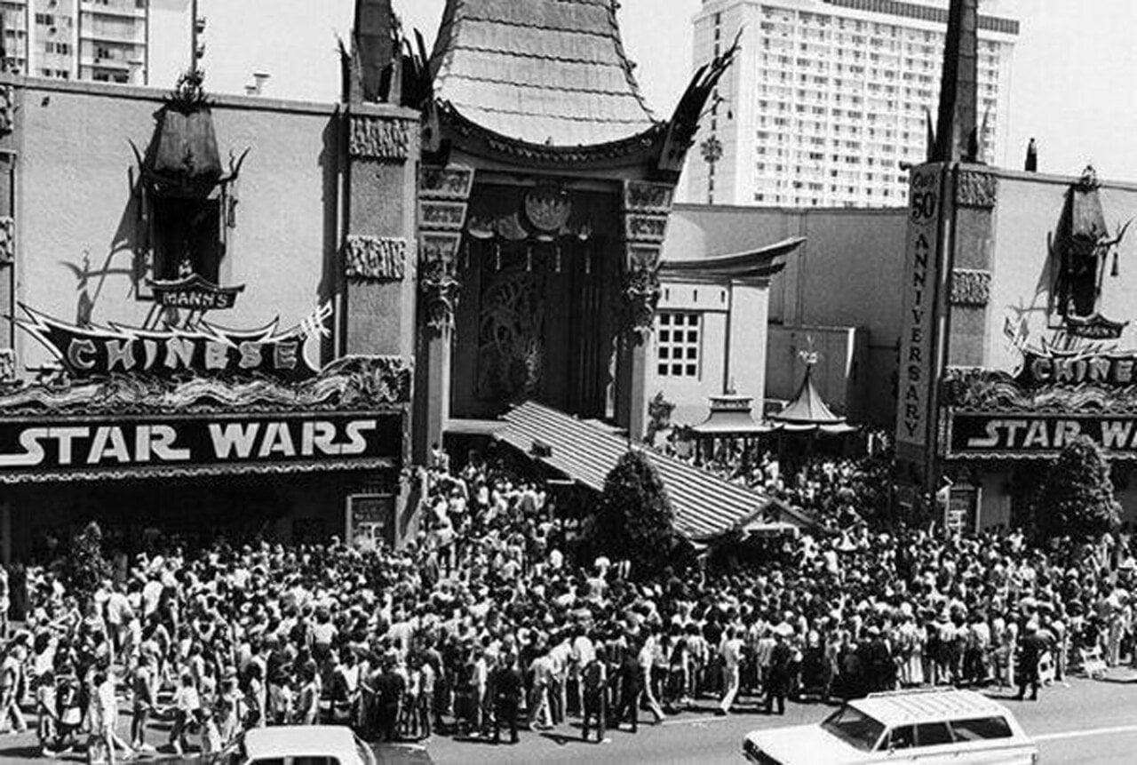 Black-and-white aerial photo of a massive crowd filling the sidewalk and street outside Mann's Chinese Theatre in Hollywood on opening day of Star Wars, May 25, 1977. The theater's marquee reads 