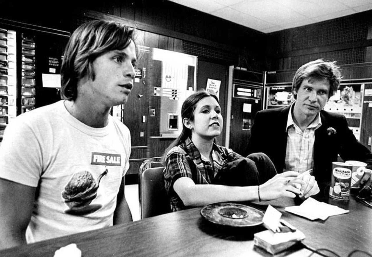 Black-and-white photo of Mark Hamill, Carrie Fisher, and Harrison Ford seated at a table during a press interview in Denver, 1977. Hamill wears a 