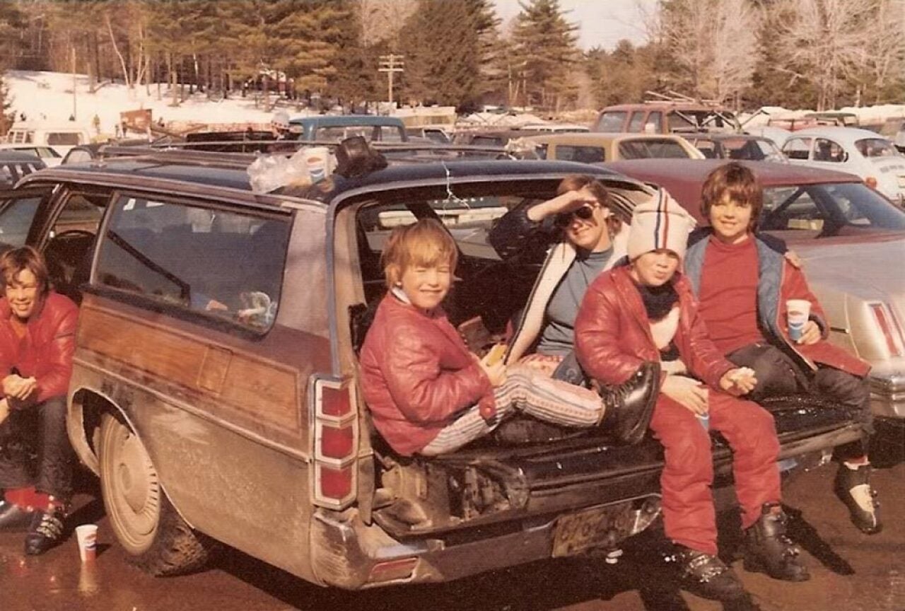 Color photo of four children and an adult sitting in the open back of a wood-paneled station wagon in a snowy ski mountain parking lot in 1977. The kids wear red ski jackets and ski boots and hold drinks and food. Pine trees and rows of parked cars are visible in the background.