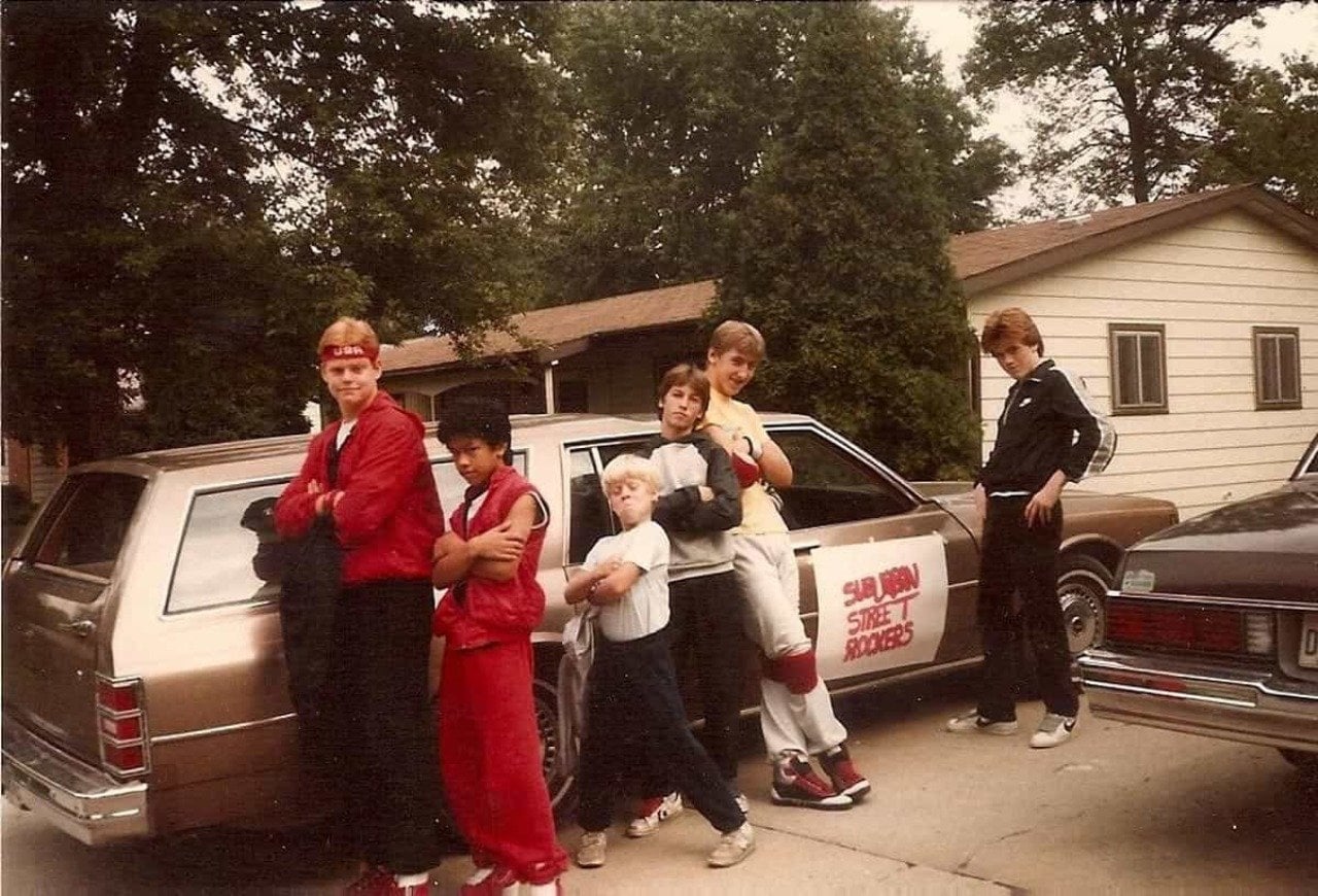 Six kids striking a pose in a driveway next to a station wagon with a sign tapped on it that reads 