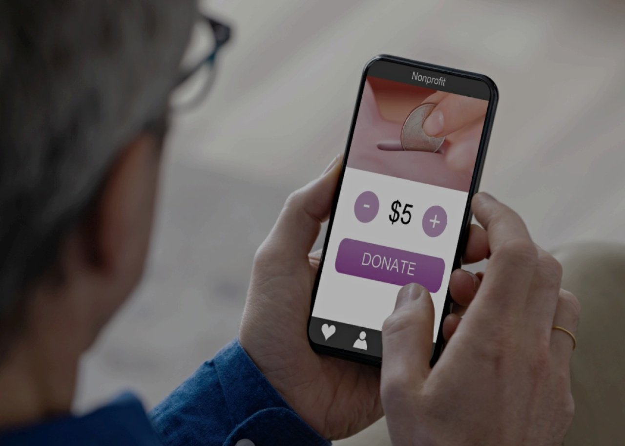 A person holding a smartphone, with a donation application on the screen displaying a coin being dropped into a slot, a selectable donation amount set to 
