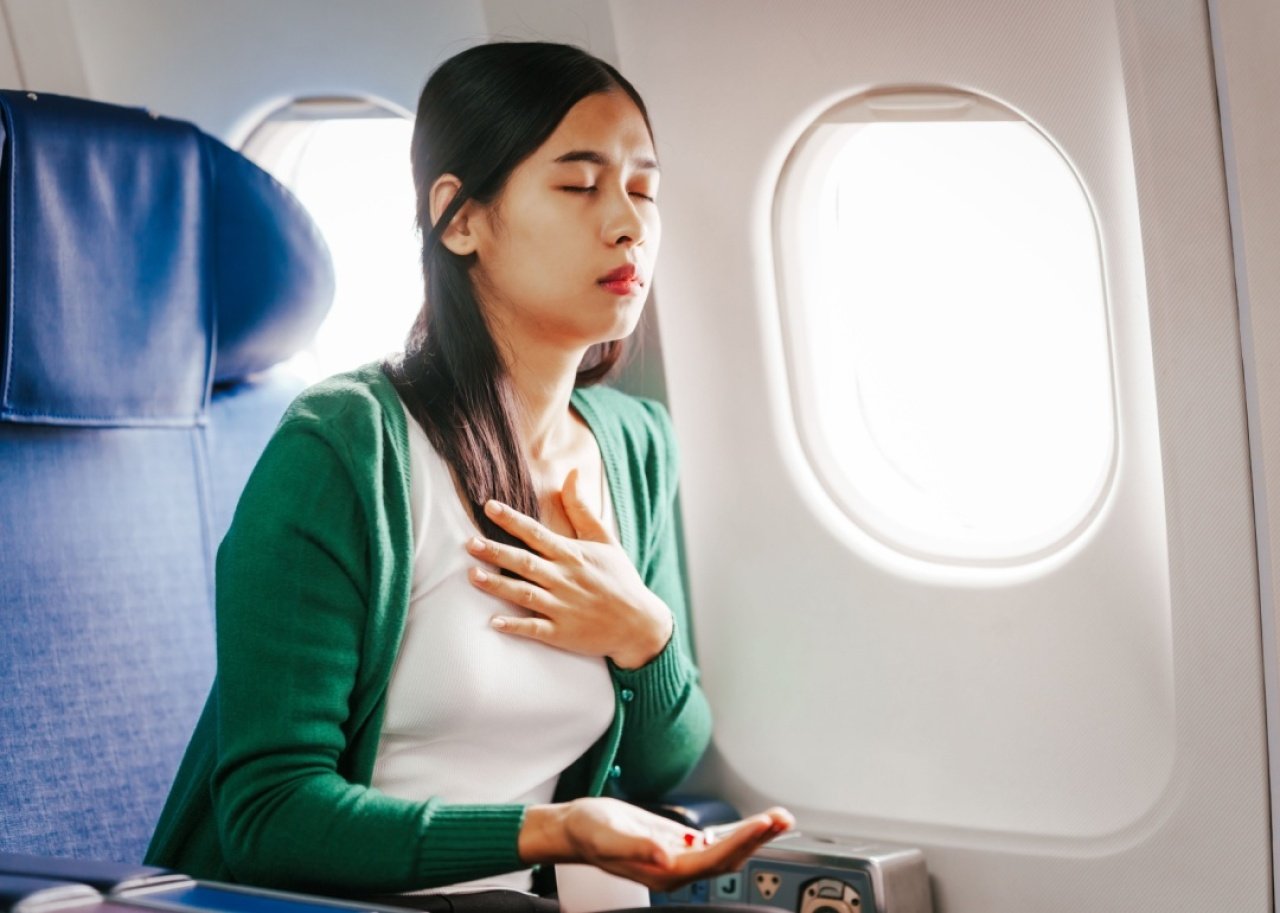 A woman is sitting in her seat as an airplane passenger, closing her eyes with her hand to her chest and other hand holding pills.