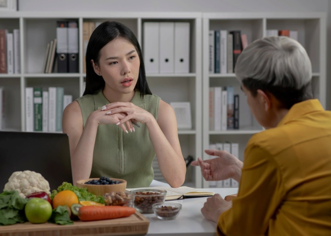 A woman looking at the older woman sitting in front of her. They are in the office with files in the background and a plate of vegetables and fruit on the desk.