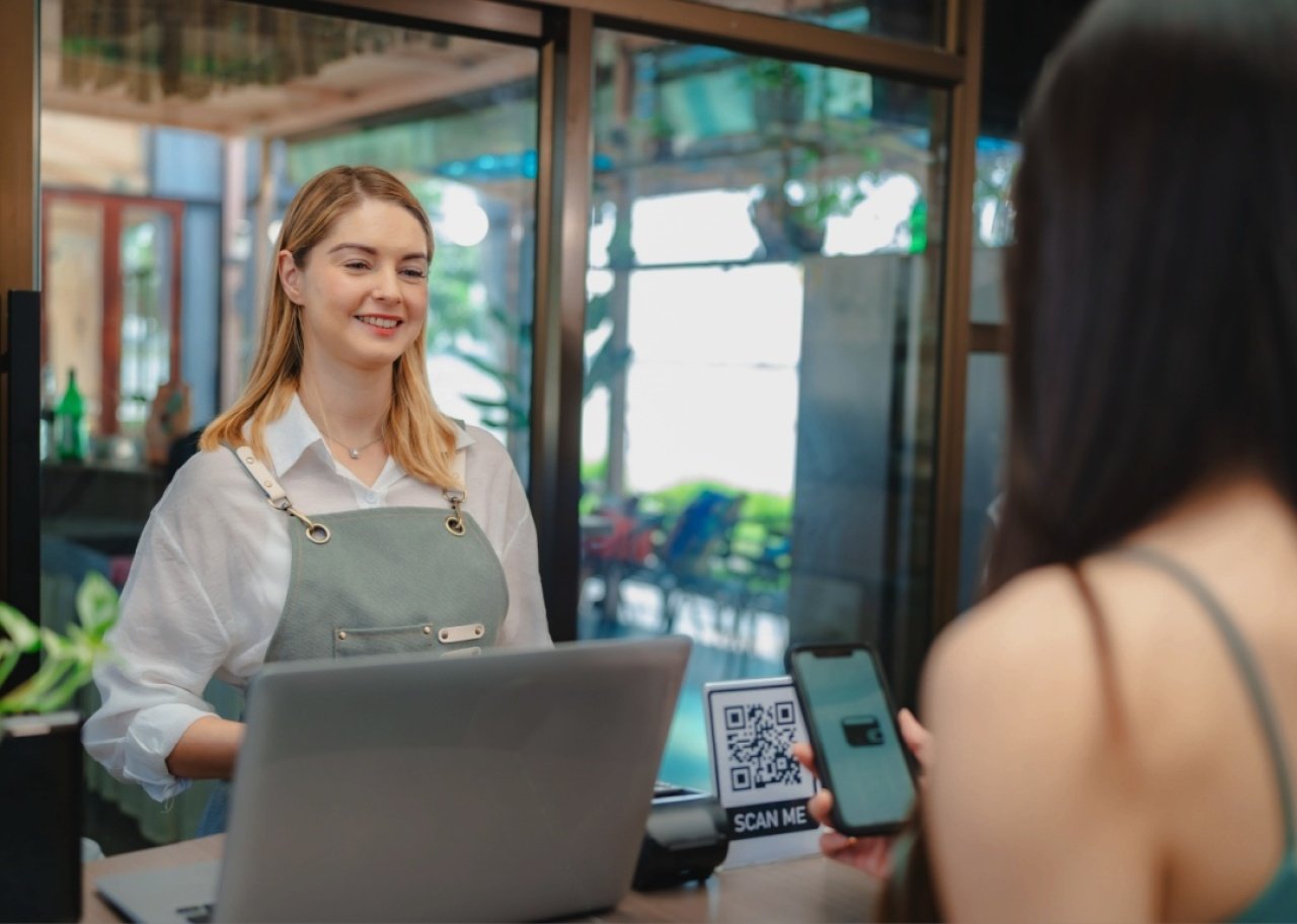 A smiling female cashier, wearing a light green apron is assisting a customer. The customer, seen from behind, is holding a smartphone and scanning a QR code displayed on the counter for payment. 