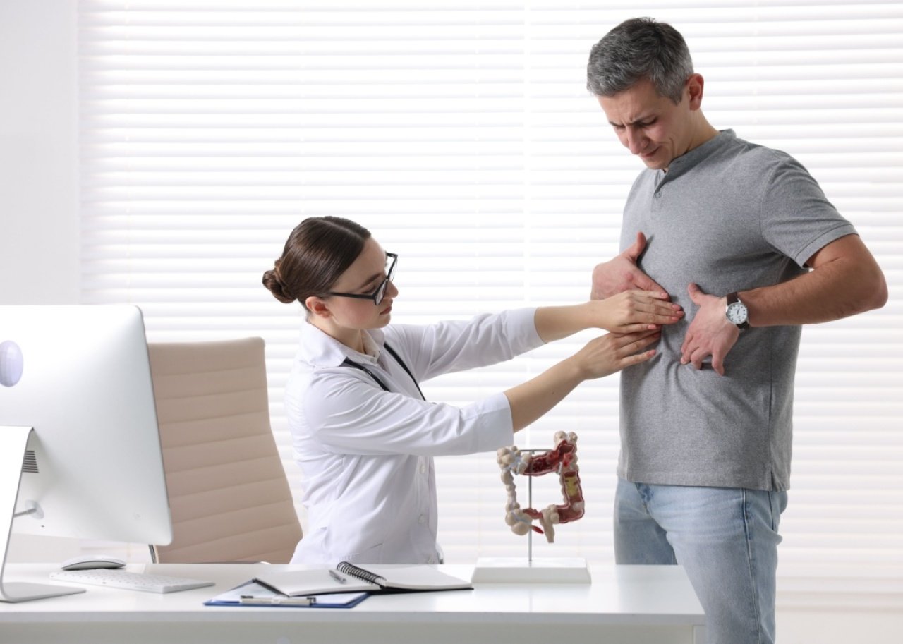 A doctor sitting at her desk is examining a patient who is holding his hands on his stomach. 