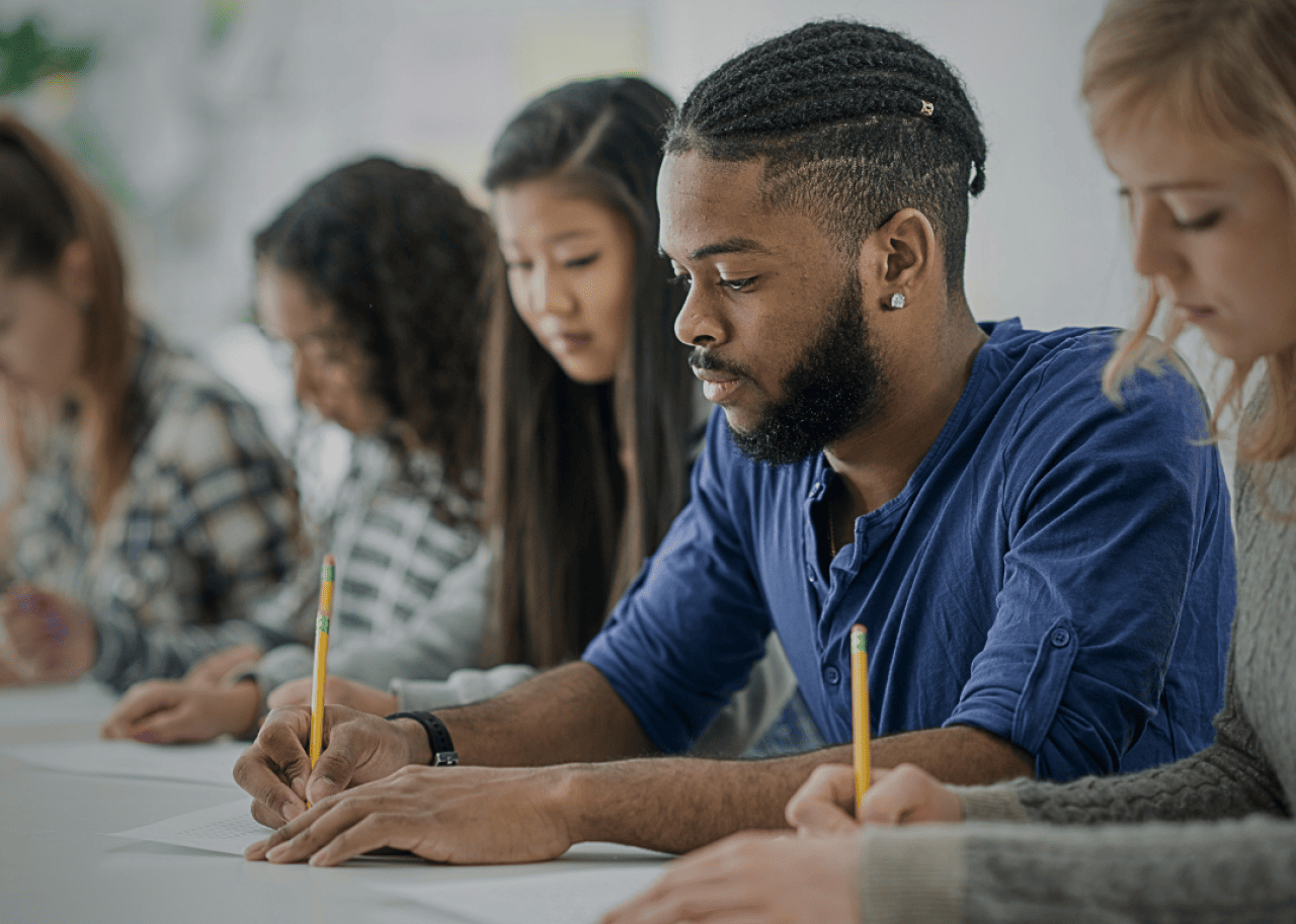 A group of students taking a test. An African American male student in the front center of the image with other female students in the background. 