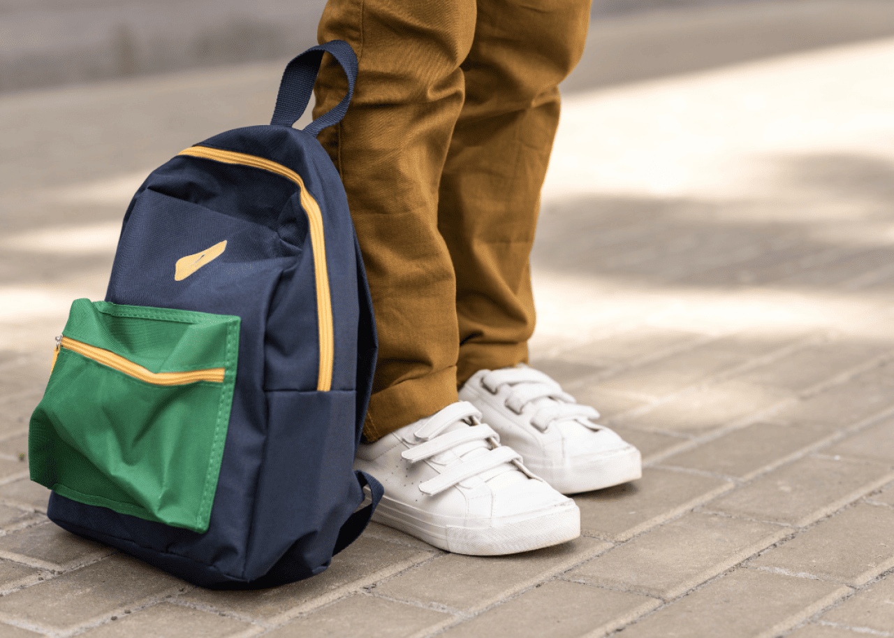 A student standing next to their backpack as seen from the knee down.