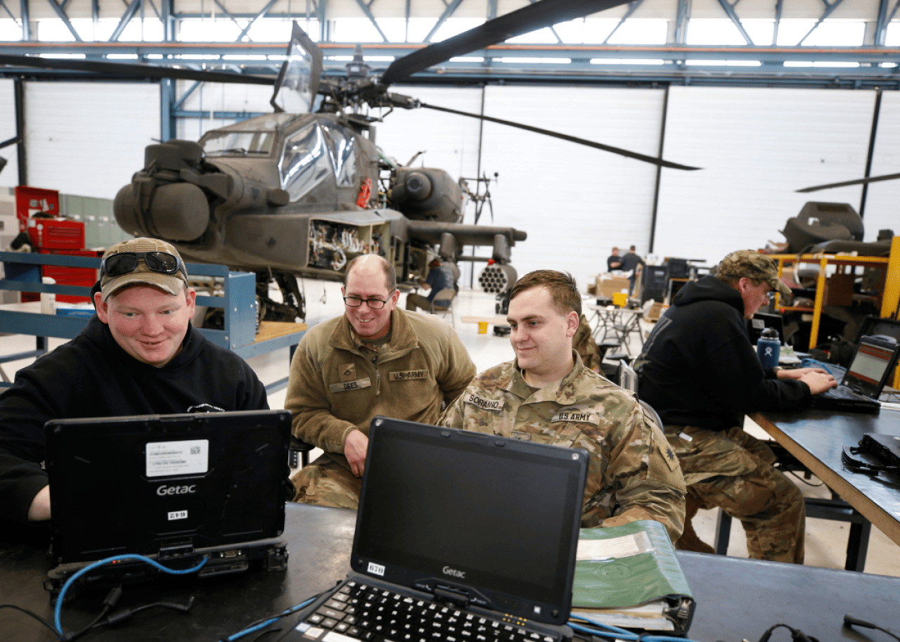 Military personnel on computers in front of a helicopter in a bright warehouse.
