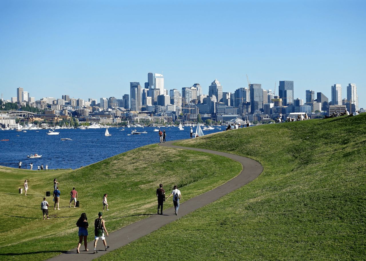 People walking in a park on the water with downtown Seattle in the background.