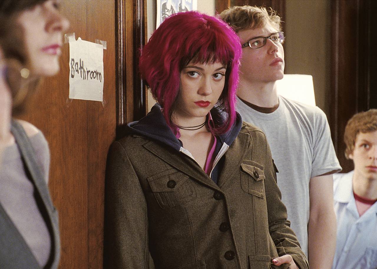 A girl with pink hair waits in a bathroom line while a boy peeks around the corner at her.