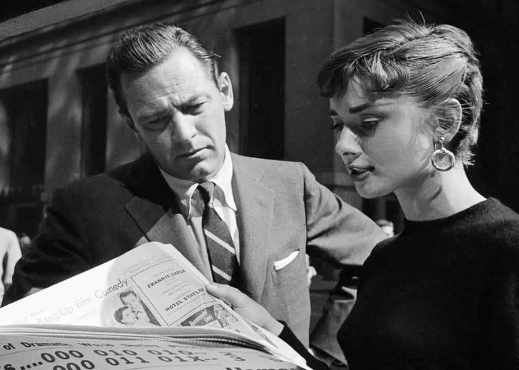 Black and white image of a woman reading a newspaper and talking to a man.