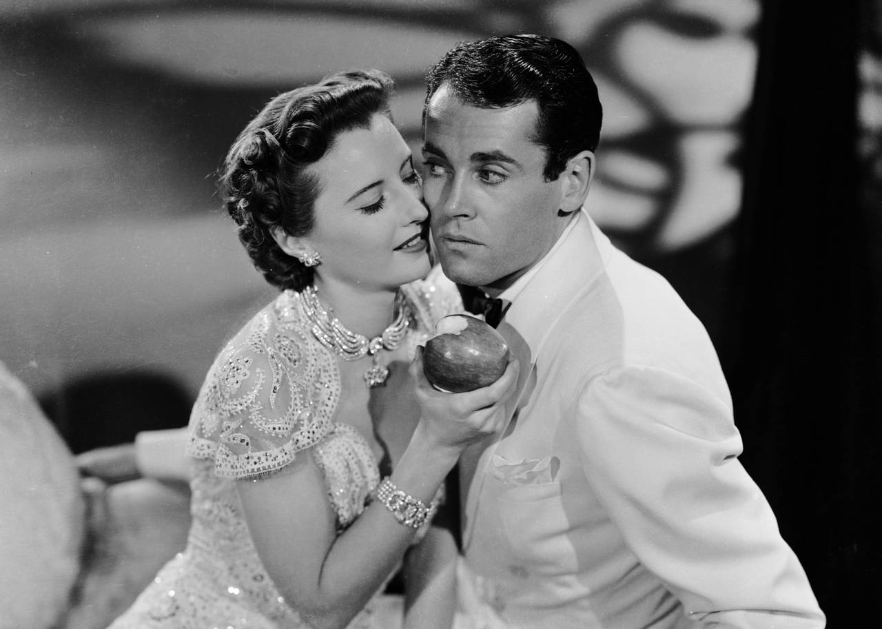 A man and woman stand cheek to cheek while she holds an apple.