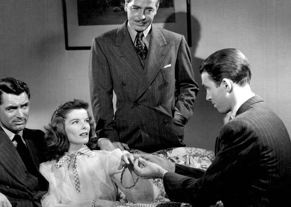 A woman, leaning back on another man, has a man putting a ring on her finger.