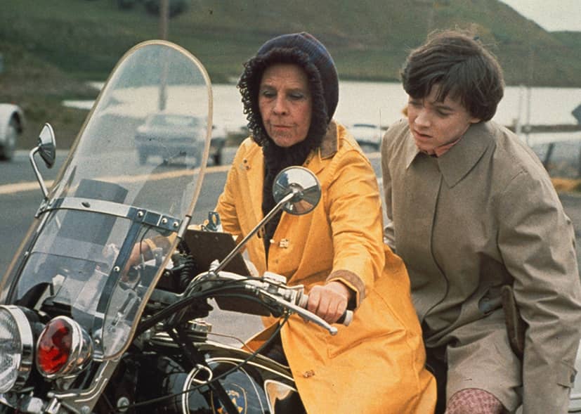 A woman in a yellow raincoat rides a motorcycle with a boy on the back.