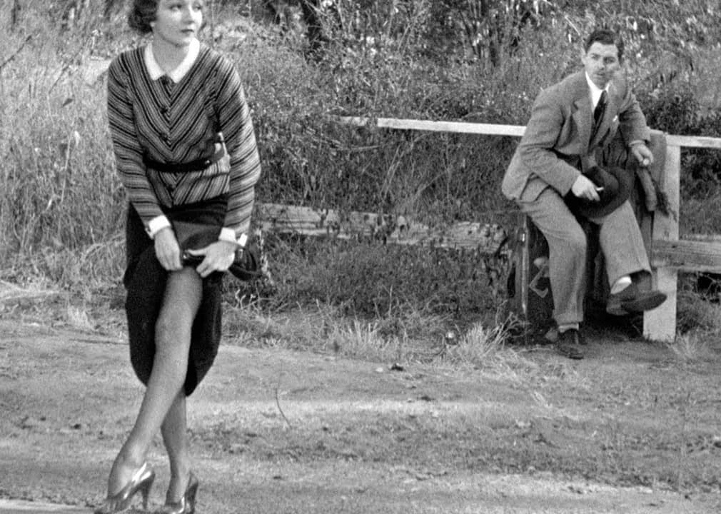 A woman adjusts her pantyhose while a man stands in the background.