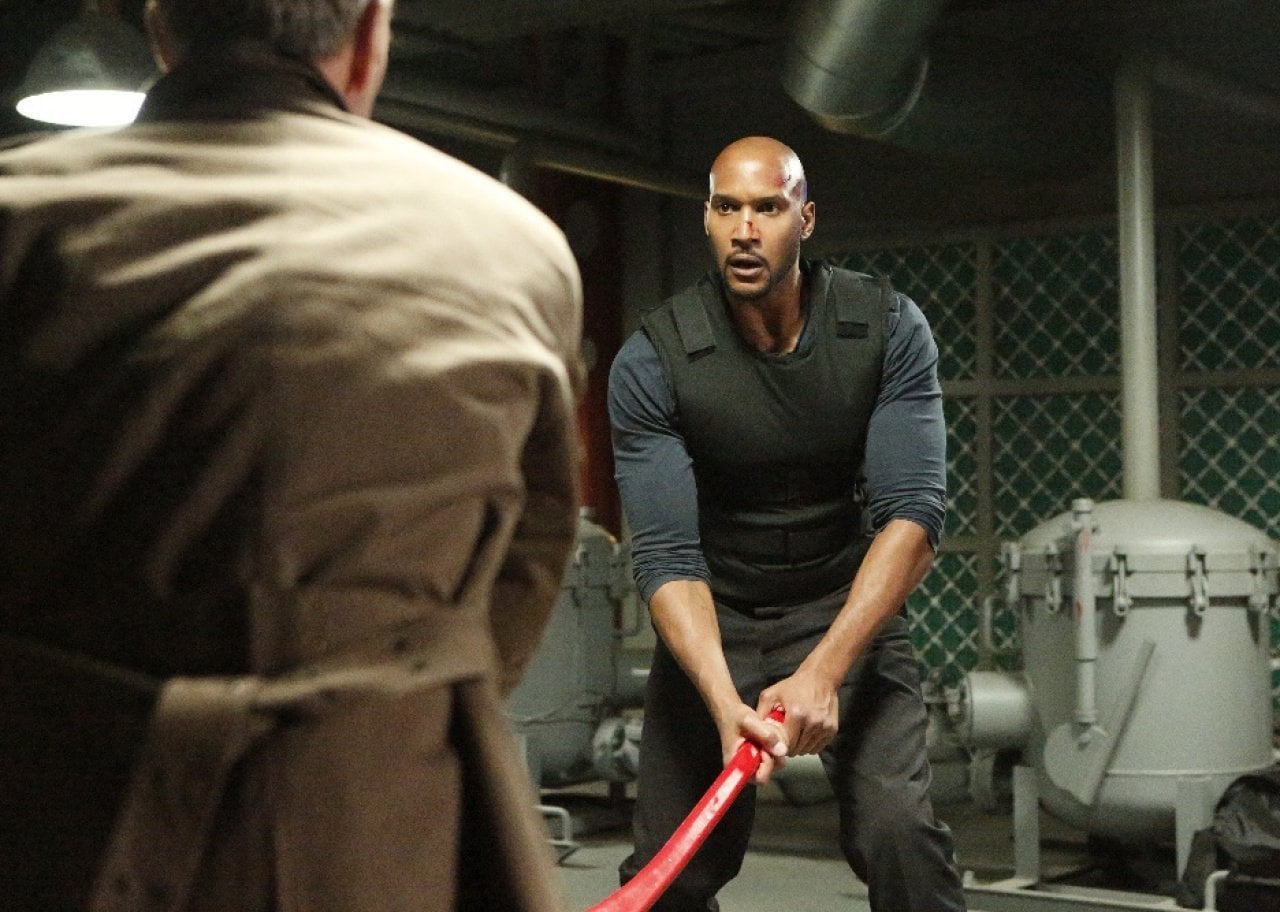 A man in a bullet proof vest holding a red axe down at the ground fights with a man in a trench coat.