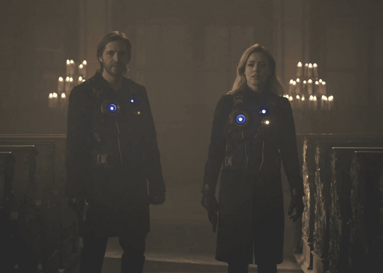 A man and woman stand with blue lights on their clothing in a dark room lit by candles.