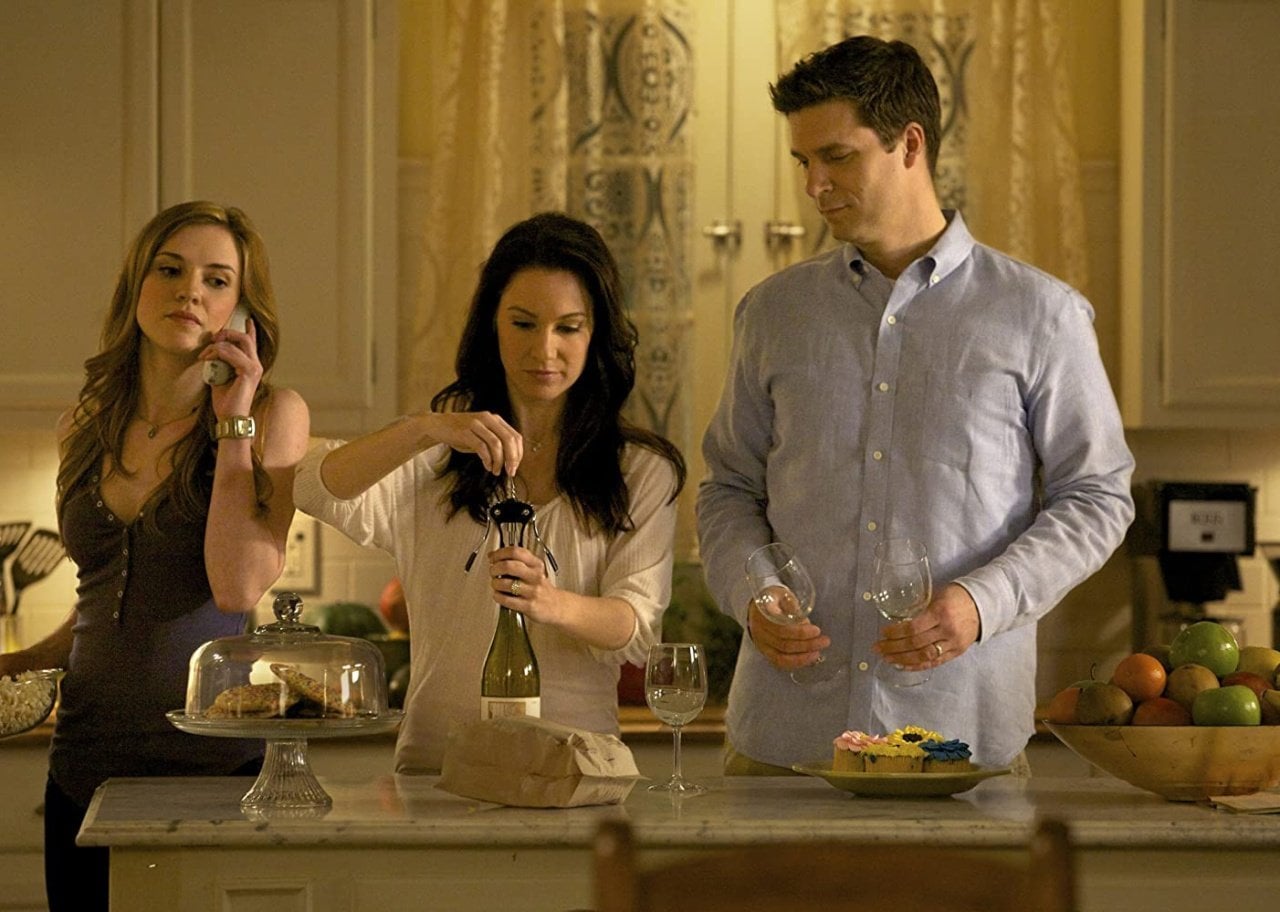 A woman opening a bottle of wine with another woman on the phone next to her and a man holding wine glasses.