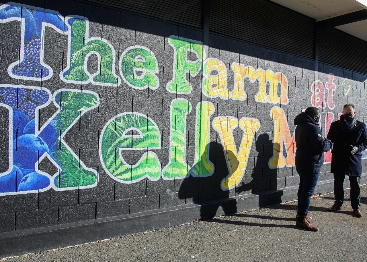 Doug Emhoff speaks with Christopher Bradshaw, executive director of Dreaming out Loud in front a brick wall that reads The Farm at Kelly Miller.