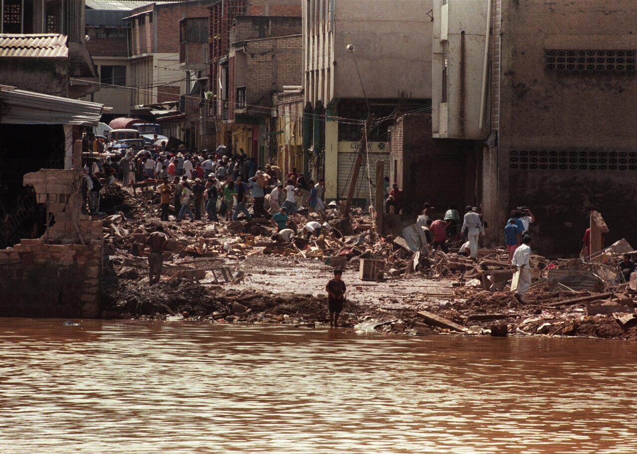 People gather in the streets of an urban area, surrounded by wreckage as the flood waters near.