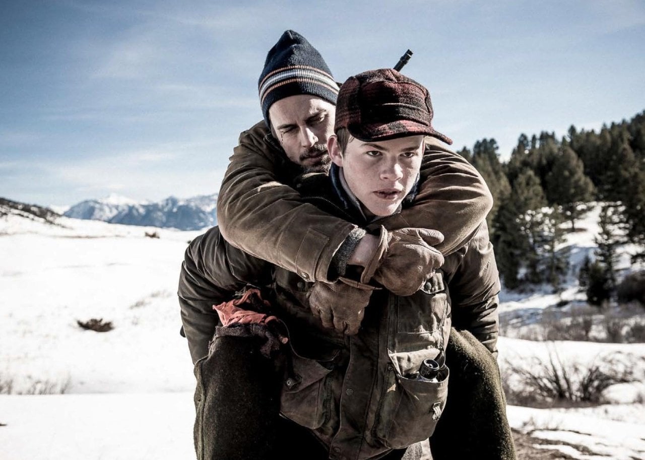 A boy carrying an injured man on his back in the snow.