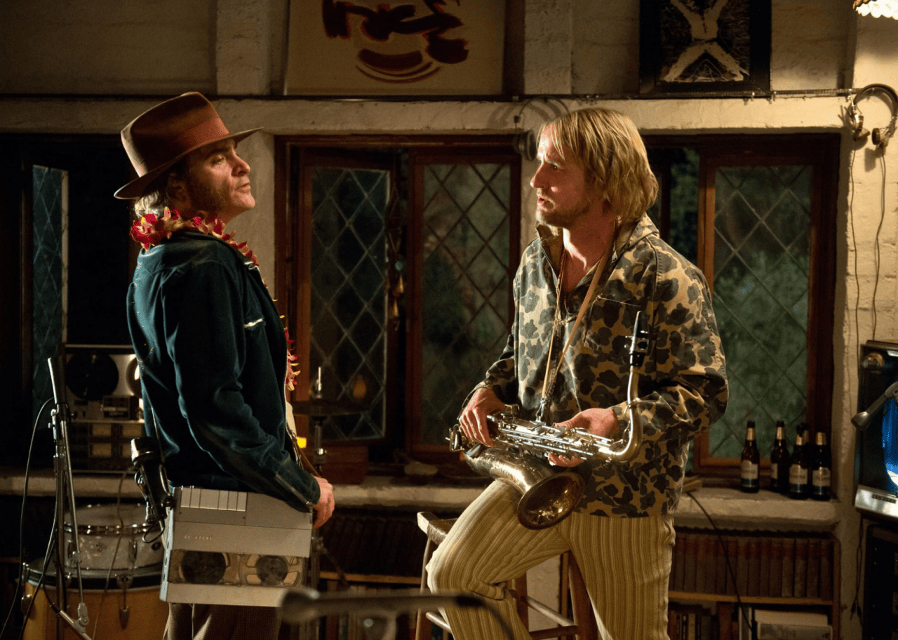 Joaquin Phoenix, wearing a recording device on a strap, standing next to Owen Wilson holding a saxophone in Inherent Vice.