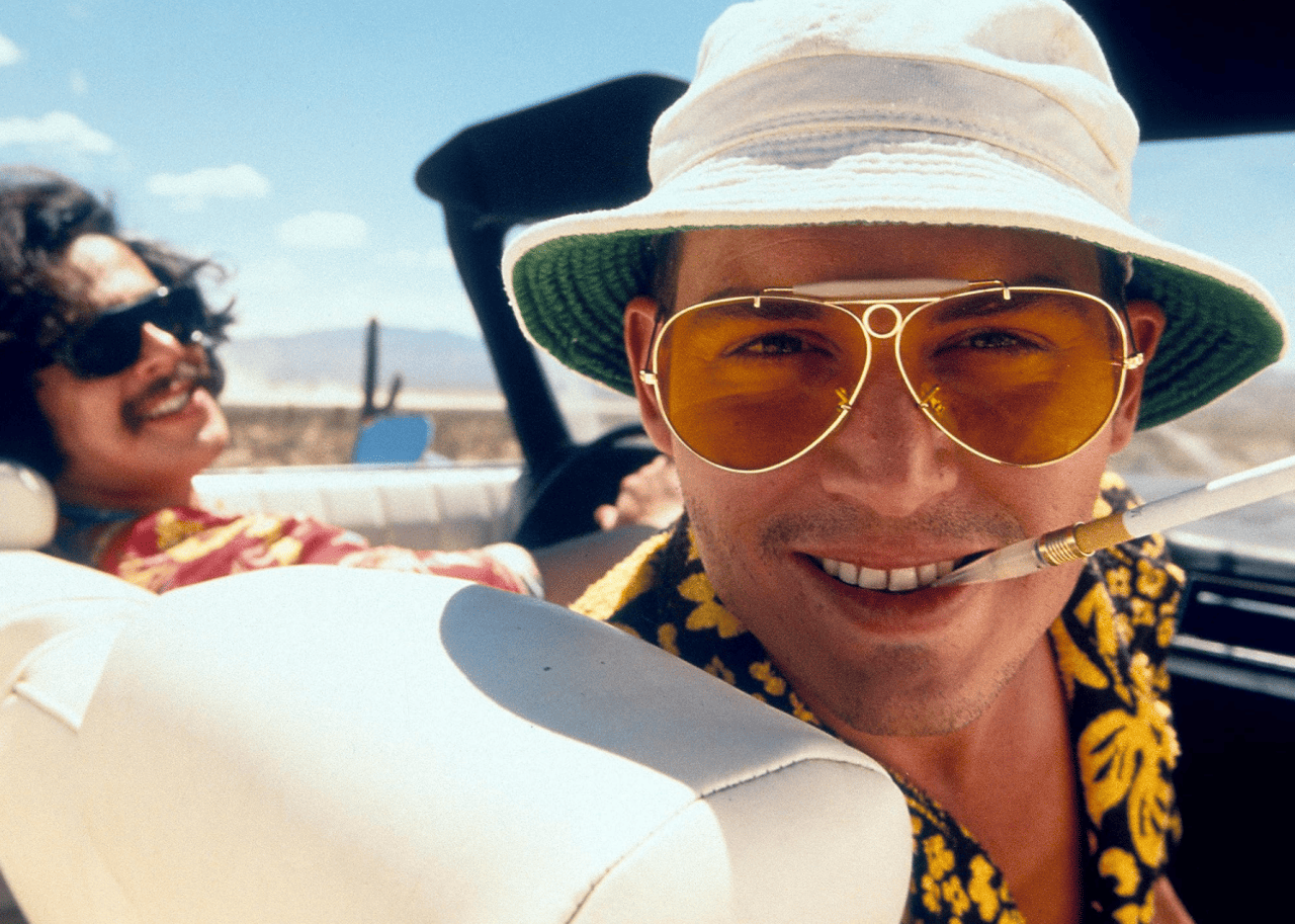 Johnny Depp with a cigarette in his mouth in the passenger seat of a convertible with Benicio Del Toro driving in a scene from Fear and Loathing in Las Vegas.