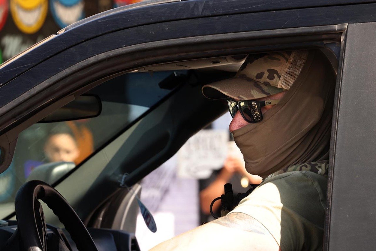 Closeup of a masked federal agent wearing sunglasses and a hat seen through his drivers' side window, planning his escape through an angry crowd after an immigrant raid on Atlantic Boulevard in the city of Bell