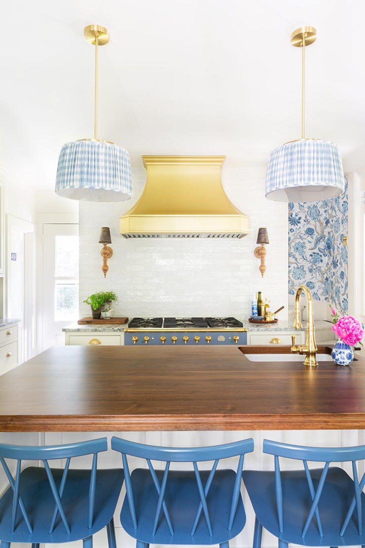 Brass range hood with white tile blacksplash, in the foreground are two hanging lamps with blue and white lampshades and an island with a wooden countertop and three blue chairs.