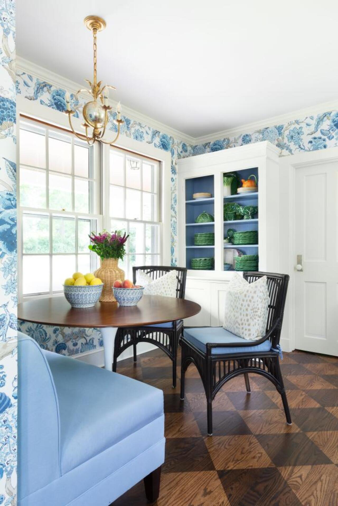 Kitchen seating area showing harlequin floor, brass hanging lighting fixture, blue upholstered chairs, blue and white wallpaper and open cabinet design in the background.