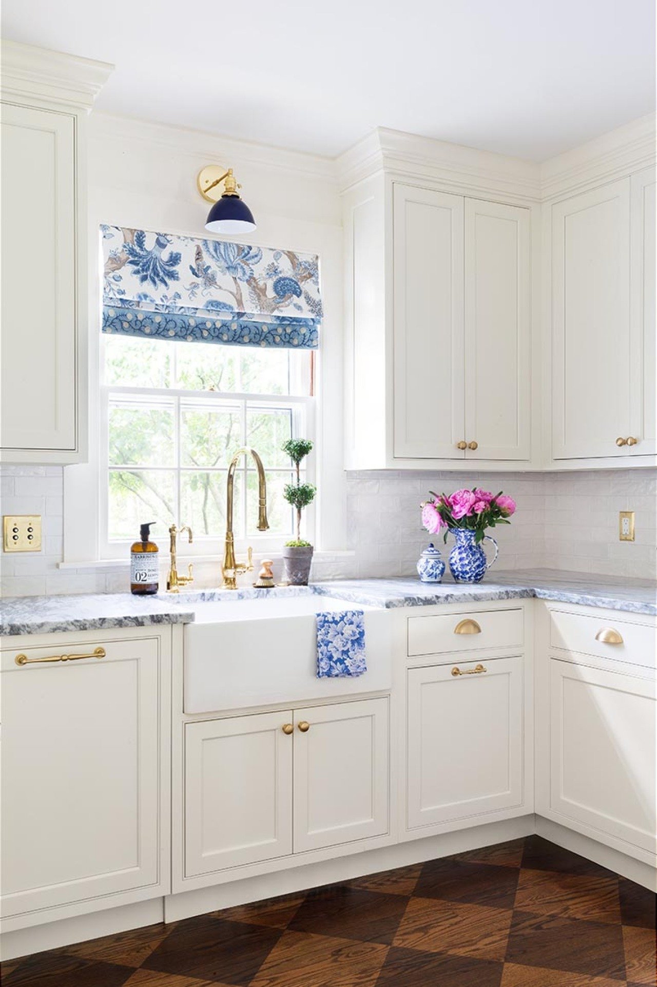 White cabinetry and a golden faucet with a window curtain that is blue and white like the wallpaper.