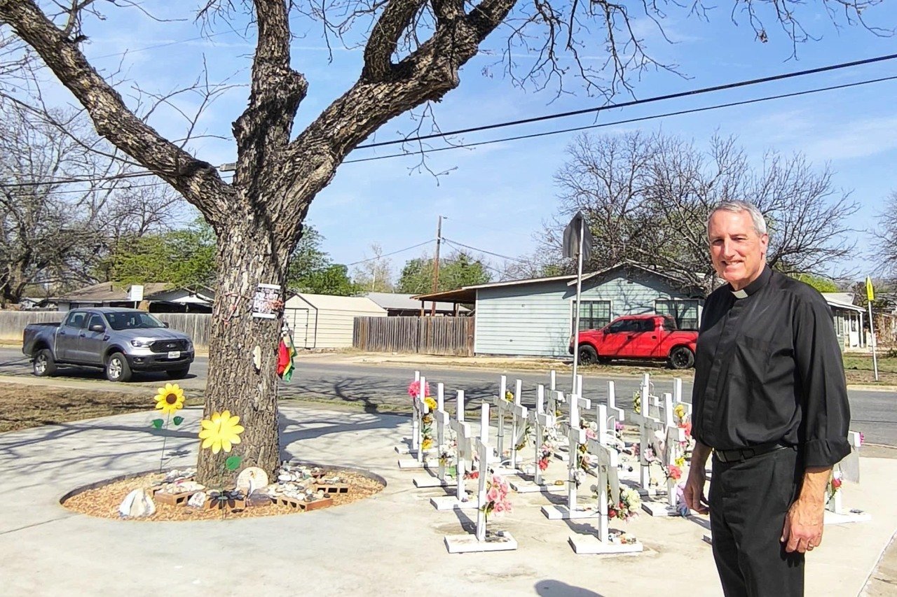 Father Michael Marsh stands in front of a memorial for the Uvalde shooting victims which is on the ground next to a tree with no leaves.
