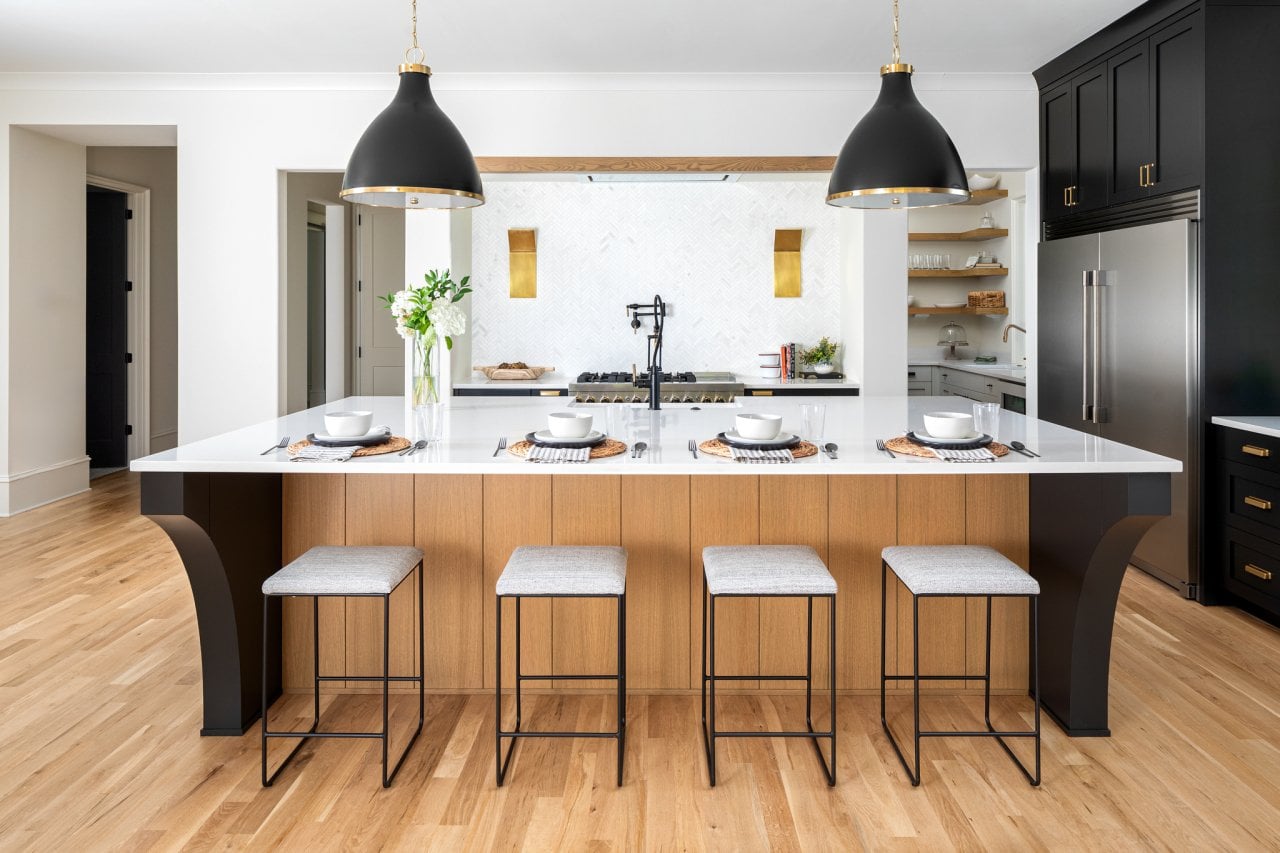 A kitchen with a white backsplash over the range, black cabinetry and light fixtures, a large chrome fridge with double doors and a center isand with four stools.