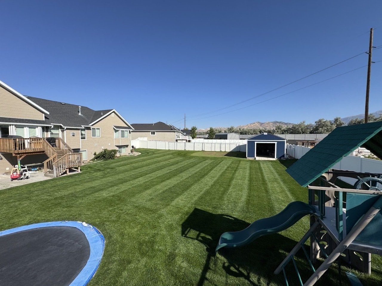 Backyard view of green lawn mowing striping patterns, with children's trampoline and jungle gym and slide in the foreground.