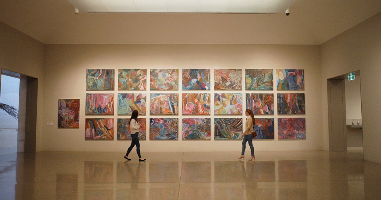 Two visitors walk by a wall where 22 canvases of paintings from