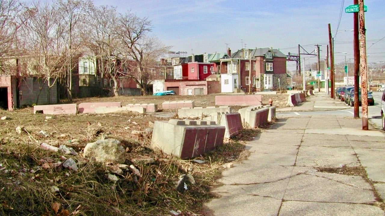View of 1600 North 54th Street before clean-up. Mostly empty lot is full of concrete fails, brown grass, litter. A row of brick homes is visible in the background.