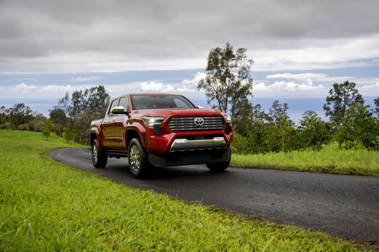 Front view of red 2026 Toyota Tacoma driving on an asphalt road with green grass on either side and gray clouds in the sky.