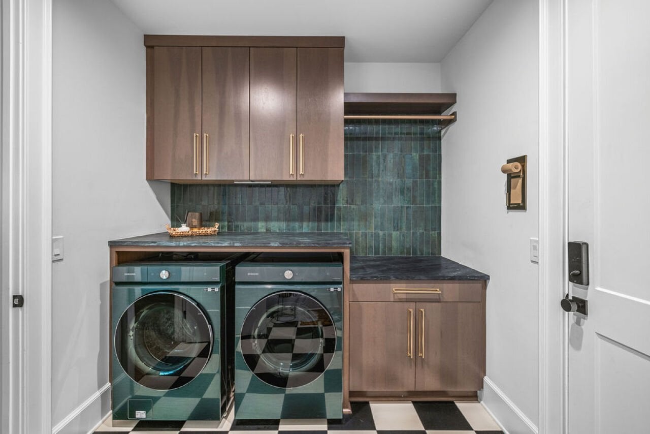 Modern laundry room with dark turquoise appliances, tiled backsplash, wooden cabinets and with brass hardware and a black-and-white checkerboard floor.