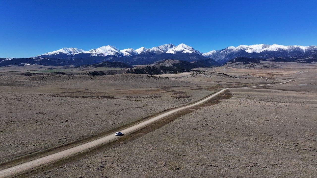 Aerial photo of single car driving on a long dirt road heading toward a mountain range, the Crazies. They resemble a mountain fortress — an island of jagged peaks rising more than 7,000 feet above the surrounding high plains, complete with secluded river valleys and alpine lakes.