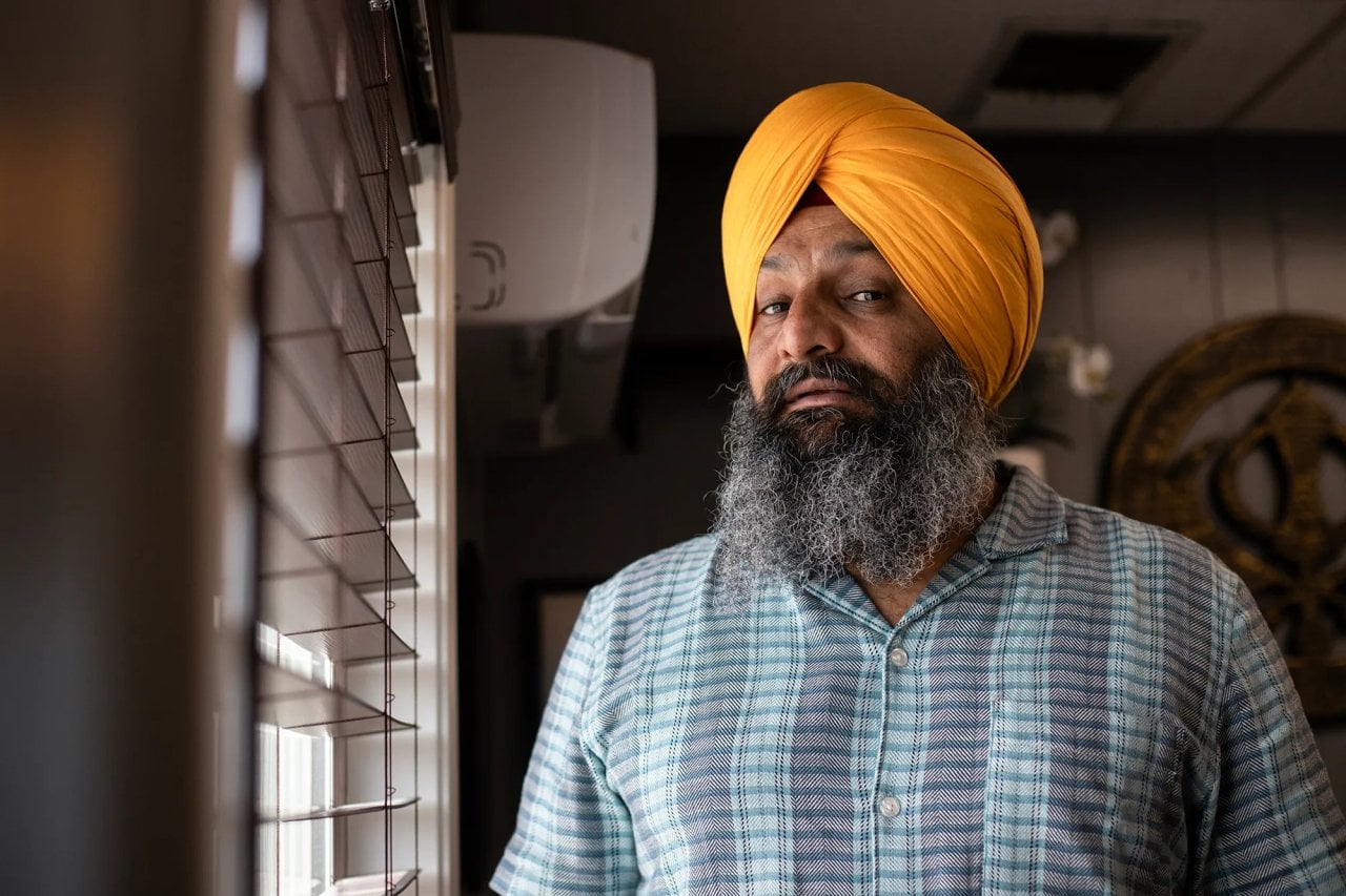 Harsimran Singh, who is president of the American Kabaddi Federation has a graying beard and a bright orange turban and he stands indoors beside a window with horizontal blinds. Soft light from the window falls across his face, creating gentle shadows. He wears a short-sleeve patterned button-up shirt and look toward the camera with a calm, steady expression. In the background, a circular wall decoration and parts of the room’s interior are visible.