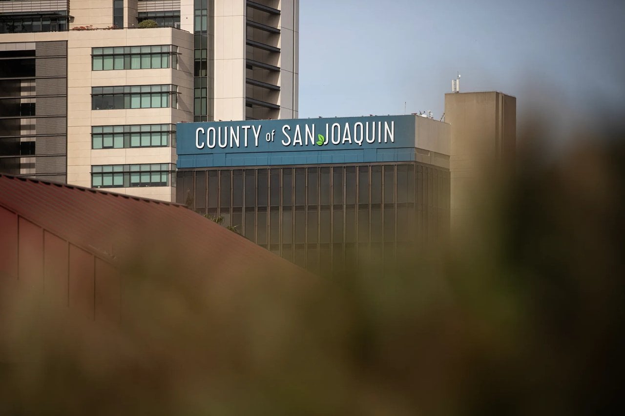 A mid-rise government building displays the words “COUNTY of SAN JOAQUIN” along its upper facade. The structure features vertical window panels and a blue band near the roofline, with taller office buildings rising behind it. Out-of-focus foliage in the foreground partially frames the lower portion of the image, adding depth.